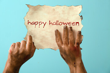 Hands of zombie holding paper with text HAPPY HALLOWEEN on light blue background