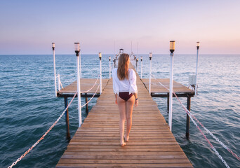 Happy young woman in bikini walking on wooden pier on the sea at sunset in summer. Lifestyle and...
