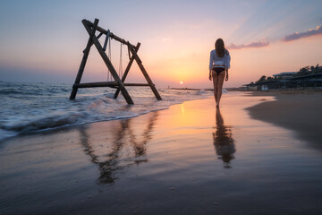 Young woman near wooden swing in water, beautiful blue sea with waves, sandy beach, reflection in water, golden sky at sunset. Summer in Side, Turkey. Girl on the sea coast. Travel. Tropical landscape