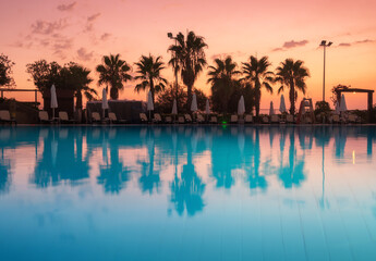 Fototapeta na wymiar Beautiful reflection in swimming pool at colorful sunset. Purple sky reflected in water, palm trees, sun beds, umbrellas at night in summer. Luxury resort. Landscape with empty pool in twilight
