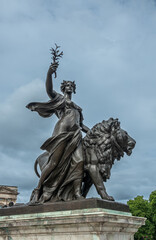 Obraz premium London, England, UK - July 6, 2022: Victoria Memorial. Closeup of Black bronze Peace statue of lion, woman holding olive branch under blue cloudscape. Some green foliage