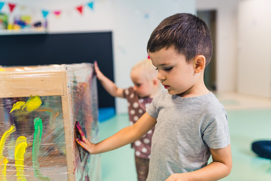 Cling wrap painting for improving kids imagination and brain development. Toddlers finger painting with tempera paints on a cling film wrapped around the wooden shelving stand. Fun activity for kids