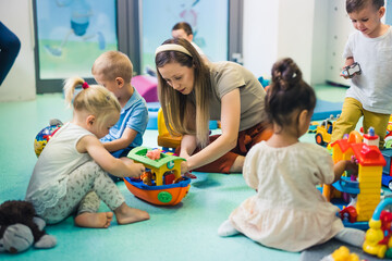 Toddlers and their tutor playing with different colorful plastic toys, cars and boats while sitting on the floor in a playroom at the nursery school. Imagination, mathematical ability, fine motor and