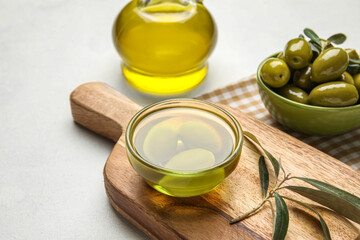 Wooden board with bowl of oil and olives on light background
