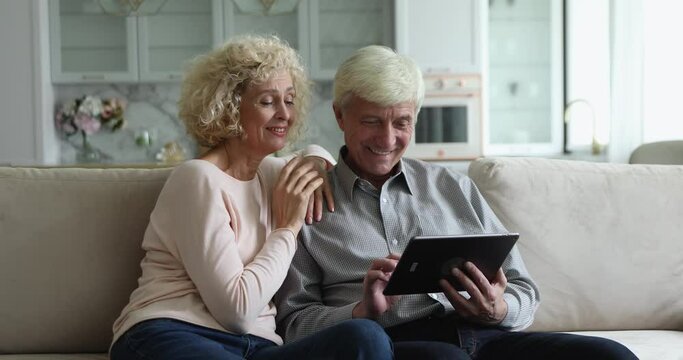 Cheerful elderly wife and grey haired husband using, discussing internet app on tablet computer, talking, laughing, smiling, shopping online, pointing at screen. Older age, modern technology concept