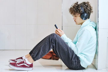 Side view portrait of depressed teen schoolboy sitting on floor and using smartphone with headphones