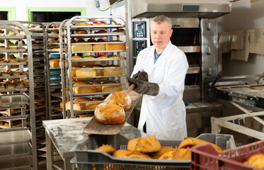 Portrait of focused man engaged in breadmaking, taking out ready loaves from oven in bakeshop..