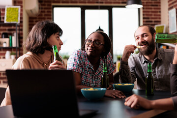 Positive diverse friends at home laughing and talking together while watching movies on laptop. Happy smiling people in living room enjoying fun leisure activity while having snacks.