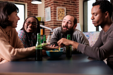 Positive group of multiethnic friends sitting at table while discussing and having fun together. Multiracial people at home in living room enjoying snacks and beverages while talking.
