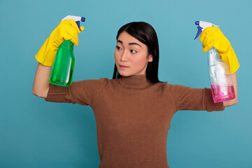 Sad tired asian housewife raising arms holding two detergent sprays in the yellow gloves against blue background, Cleaning home concept, Overworked housewife from day to day chores