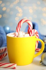 Cup of tasty cocoa with marshmallows and Christmas candy canes on white table against blurred festive lights