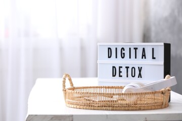 Gadgets in wicker basket near lightbox with phrase DIGITAL DETOX on white table indoors. Space for...