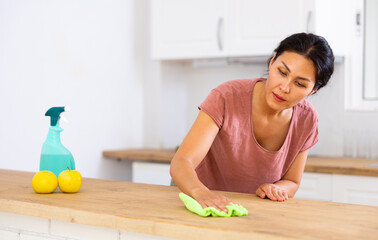 Asian woman washing top of kitchen table during cleanup in apartment.