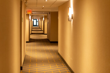A long narrow hallway of a hotel with a red exit sign light sconce lights and cream colored walls....