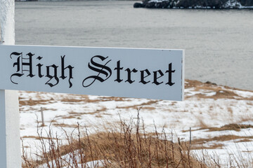 A white wooden street marker on a single wooden post. The name of the sign is High Street. The background is snow, yellow grass, an ocean, and a cliff. The lettering on the sign is black Gothic text.