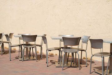 Multiple empty grey metal tables and chairs on a cafe sidewalk patio of a restaurant. The exterior brick wall and background are yellow colored with pink and gray colored tile on the ground.  