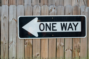 A metal directional traffic sign is attached to an unfinished wooden fence. The black and white...