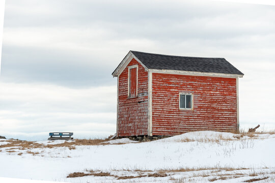 The exterior of a colorful vintage red storage building. The old wooden barn has wood siding on the walls, a small window, and multiple closed red wooden doors. Yellow grass and snow are on the ground