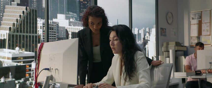 Two diverse female office employees working on something together in the modern busy office. View on a business district with skyscrapers in the background