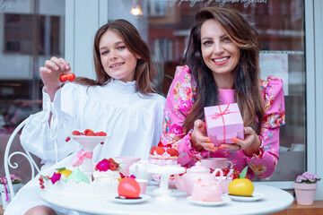 Cool satisfied smiling elegant well-dressed mother and daughter with present box, fruit, berry sweets on table in cafe