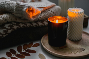 Two burning candles and stack of warm cozy sweaters on bedtable. Cozy autumn evening at home