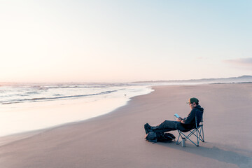 mature man sitting alone relaxed outdoors on the beach shore reading a book and listening to music at sunset