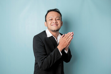 Smiling young Asian businessman wearing black suit gesturing greeting or namaste isolated over blue background
