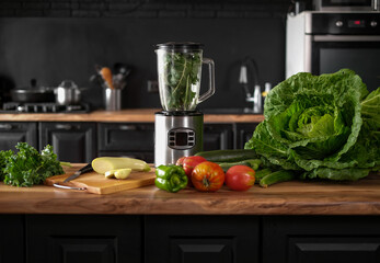 Blender full of green ingredients for smoothie making on the table with green raw vegetables on the kitchen. Black modern kitchen interior. 