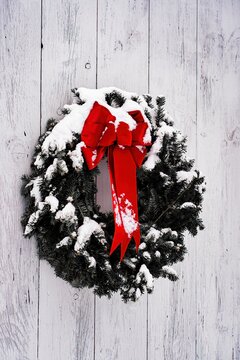 Snow-dusted pine needle Christmas wreath and red bow on whitewashed wide plank door. Rustic, traditional country Christmas. Image manipulation with near monochrome and isolated, saturated red bow.