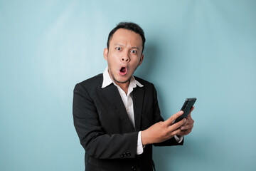 Surprised Asian businessman wearing black suit holding his smartphone, isolated by blue background