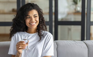 Healthy lifestyle concept. Beautiful young Afro American woman with glass of clean water, looking...