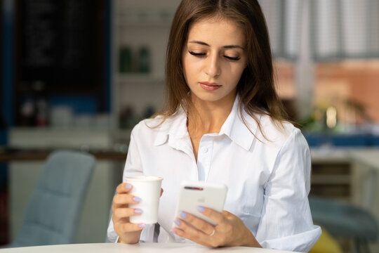Charming young woman reading good news on mobile phone while relaxing in cafe, happy girl looking at photos on cell phone in free time in coffee house