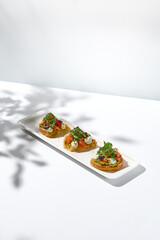 Aesthetic composition with salmon bruschetta on white background with shadows from flowers. Italian bruschetta with salmon, avocado, cheese and olives on fine dining in summer. Elegant menu concept.