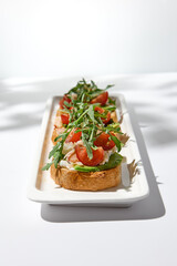 Aesthetic composition with shrimp bruschetta on white background with shadows from flowers. Italian bruschetta with shrimp, tomatoes and arugula on fine dining in summer. Elegant menu concept.
