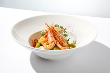 Elegant food - salad with shrimp and avocado on white table with harsh shadow. Flashy food concept. Seafood salad with shrimp, tomatoes and avocado on white plate with sunlight.