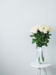 Beautiful bouquet of white blooming roses in glass vase on the white wall background. White flowers with ribbon stand on small round table. Floral still life. Copy space. Rose hybrid tea Avalanche.