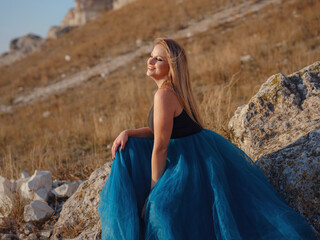 Fashionable woman on desert field near mountain wearing black top and blue tulle skirt - 525484875