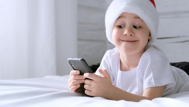 5 Year Old Boy In A Santa Claus Hat Laughing Looking Fun Video On Smartphone While Lying On Bed. Kid Have Fun Learning Using Phone. The Child Is Practicing A Foreign Language By Talking With Friends.