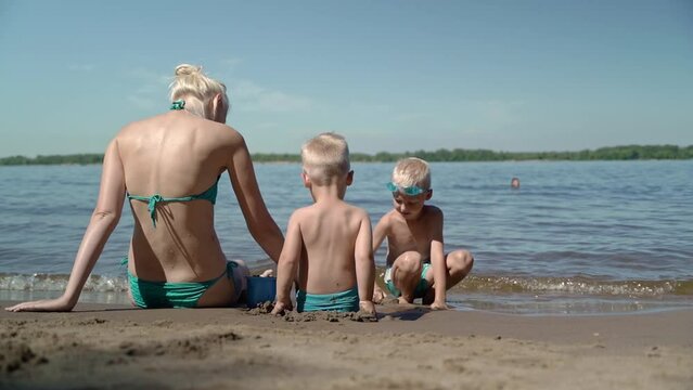 A young woman with her two sons is having a great vacation on the beach. Children are happy to be together and play with their mother. Mom shows them what figures can be made from sand.