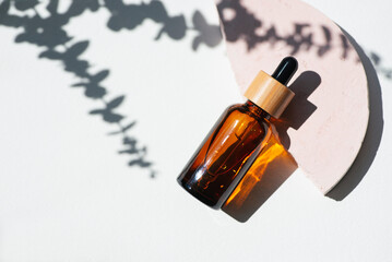Amber bottle with dropper pipette with serum or essential oil on pink concrete podium with eucalyptus shadows. White background with daylight. Beauty concept for face and body care