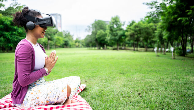 African Female woman wearing virtual reality glasses meditating in lotus position at park,yoga namaste enjoying simulation and relax in metaverse world.AR,VR Virtual Future technology concept.