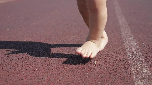 Small child legs walk on track for running competition on stadium. Barefoot baby boy imagines himself big runner in sports. Legs cast shadow on ground