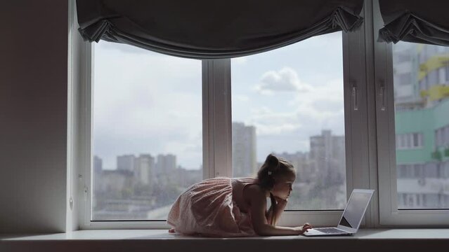 Attractive small caucasian child using laptop computer surfing or watching cartoons on a window sill at home. E-learning concept. High quality 4k footage
