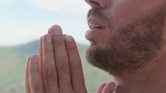 Closeup shot of a young man prays to God with his eyes closed, holding his praying hands at chin level, saying prayers and enjoying serenity and tranquility, alone against the backdrop of mountains