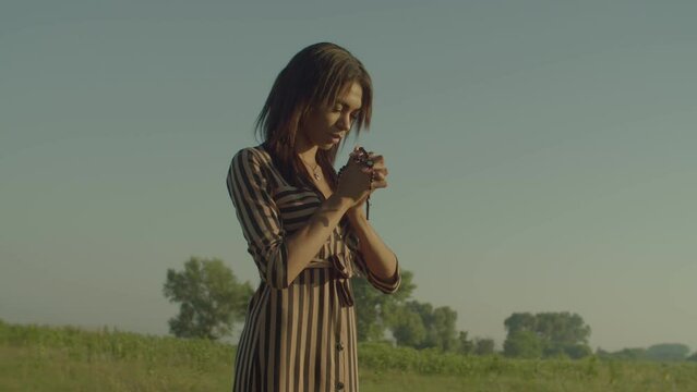 Portrait of Christian beautiful African American woman with eye closed holding catholic rosary, saying prayers, asking for bless, mercy and peace while standing on green field in morning