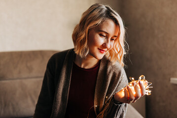 Young woman preparing for christmas and holding garland with lights. Xmas spirit in cozy modern home indoors. Portrait of people waiting holidays