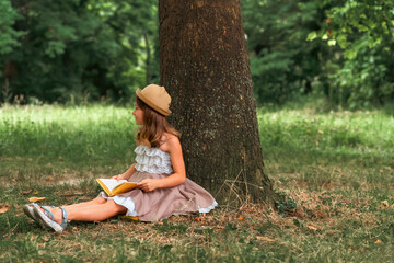 School holidays. Pretty schoolgirl girl in a straw hat and dress is sitting on the ground by a tree and reading a book. Copy space. The concept of independent extracurricular children's education