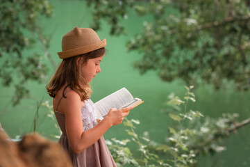 Children's education. Schoolgirl in a dress and a straw hat reading a book at the park. In the background are trees and green river. Copy space. The concept of school holidays and curiosity