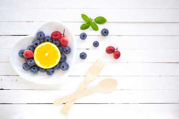 Naklejka na ściany i meble Top view mix berry,orange on white heart shape plate with wooden fork and spoon on plank white wood background.