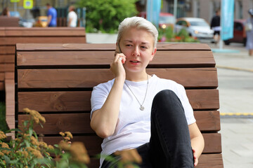 Girl with short blonde hair talking on mobile phone while sitting on street wooden bench. Tomboy...